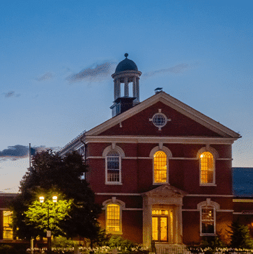 Historic red-brick building with lit windows and a cupola at dusk.