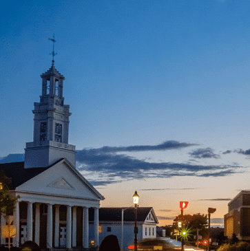 Small town street with a white church and a tall steeple under a twilight sky.