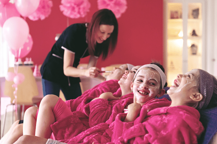 Children enjoying a spa day with facials, wearing pink robes and headbands while smiling.