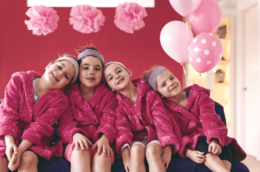 Four smiling children in pink robes and headbands, sitting together with pink balloons behind them.