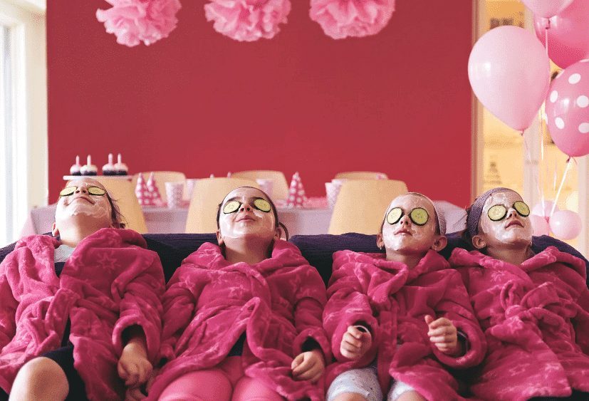 Four girls in pink robes enjoy a spa day with face masks and cucumber slices on their eyes.