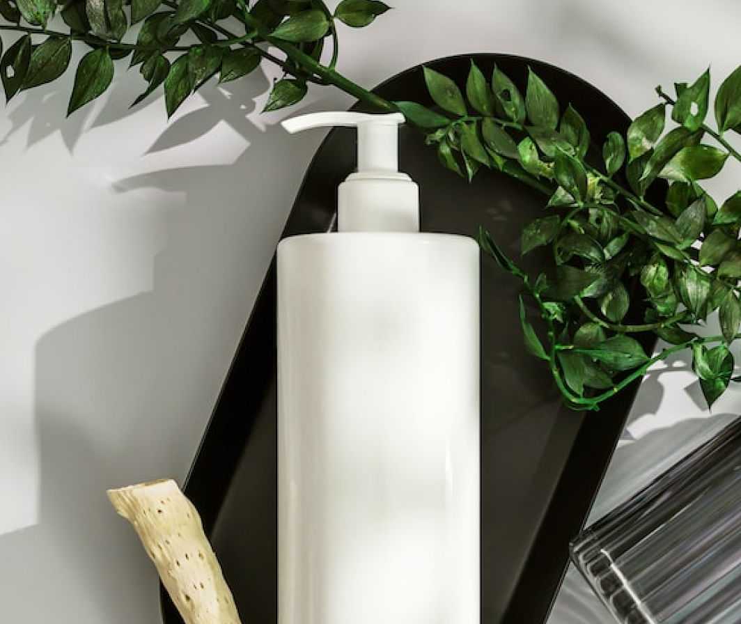 White lotion bottle with pump surrounded by green leaves on a black tray, with sunlight casting shadows.