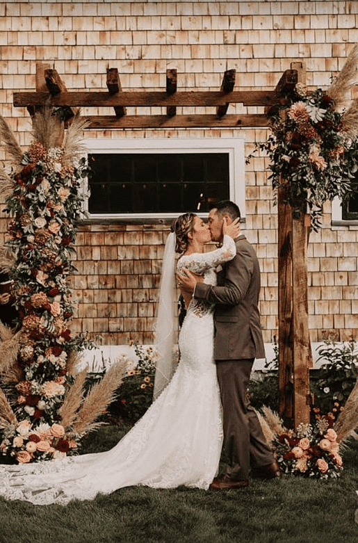 Bride and groom kissing under rustic wooden arch adorned with flowers and greenery.